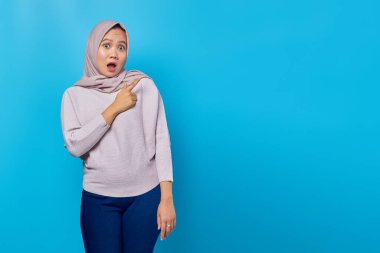 Şok olmuş Asyalı kadın portresi. Parmağıyla boşluğu işaret ediyor. Mavi arka planda ağzı açık.