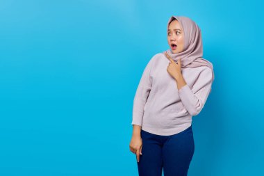 Şok olmuş Asyalı kadın portresi. Parmağıyla boşluğu işaret ediyor. Mavi arka planda ağzı açık.