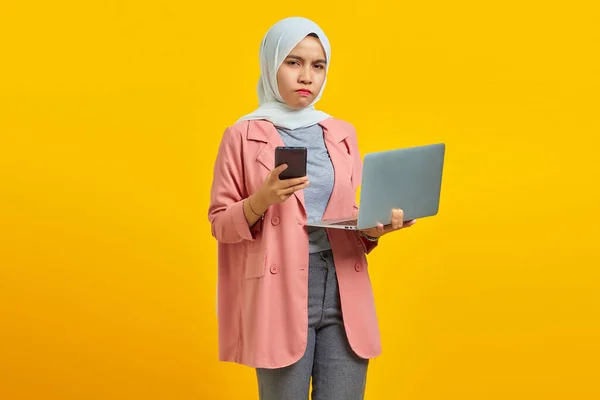 在蓝色背景下拿着手机和笔记本电脑的愤怒而不快乐的年轻亚洲女性 — 图库照片