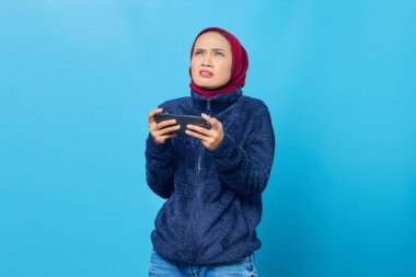 Şok olmuş genç Asyalı kadın akıllı telefondan video oyunu oynuyor ve mavi arka planda