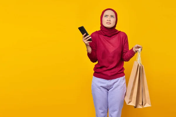 Töprengő Fiatal Ázsiai Kezében Mobiltelefon Bevásárló Táskák Sárga Alapon — Stock Fotó
