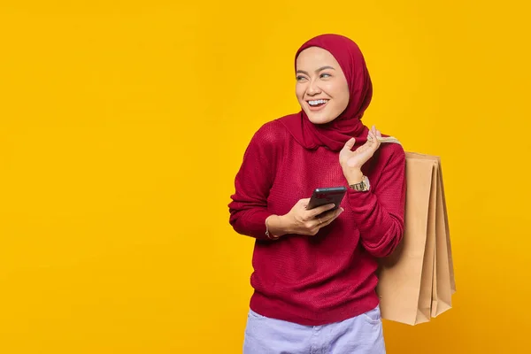年轻而快乐的亚洲女人一边拿着购物袋和智能手机 一边看着黄色的背景 — 图库照片