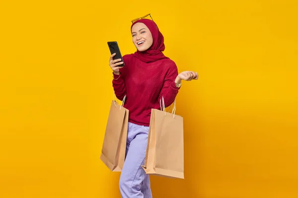 携帯電話でアジアの若い女性を笑顔と黄色の背景にショッピングバッグを保持 — ストック写真