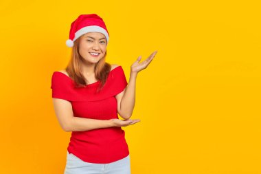 Noel şapkası takan güzel Asyalı bir kızın portresi sarı arka planda avuç içi izole boşluğu işaret ediyor.