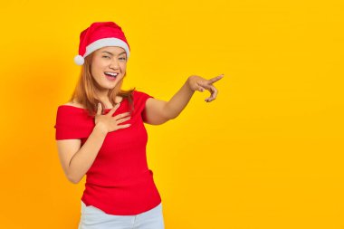 Noel şapkası takan neşeli genç Asyalı kadın sarı arka planda kamerayı işaret ediyor.