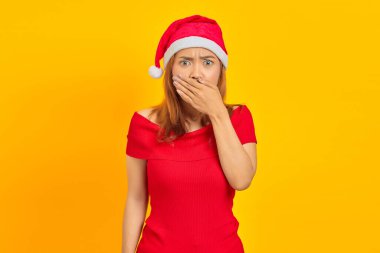 Şaşırmış genç Asyalı kadın Noel şapkası takıyor ağzını örtüyor sarı arka planda elleri var