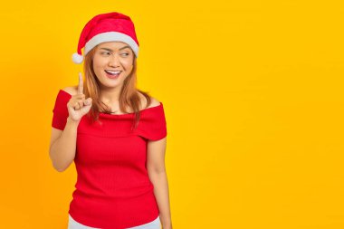 Gülümseyen Asyalı genç bir kadın Noel şapkası takıyor parmaklarını havaya kaldırıyor ve sarı arka plan hakkında bir fikri var.