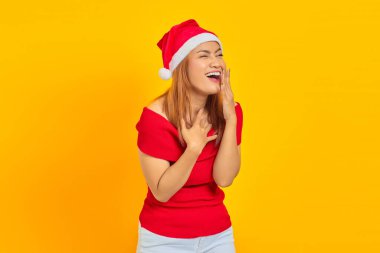 Genç Asyalı kadın portresi Noel şapkası takıyor elleriyle ağzını kapatırken gülen yüz ifadesi.