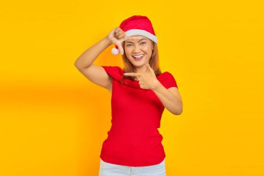 Sarı arka planda parmakları izole bir çerçeve yaparken Noel şapkası takan neşeli Asyalı genç bir kadının portresi