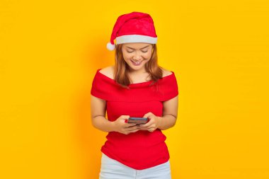 Sarı arka planda akıllı telefondan mesaj gönderen, Noel şapkası takan gülümseyen genç bir kadının portresi.
