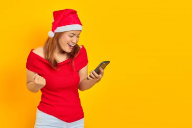 Noel şapkası takan neşeli genç Asyalı kadın cep telefonuyla sarı arka planda başarıyı kutluyor.