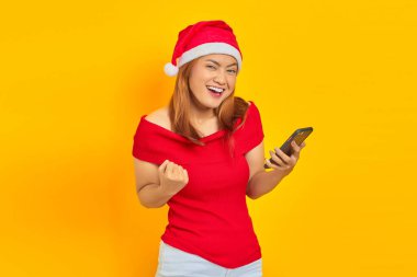 Noel şapkası takan neşeli genç Asyalı kadın cep telefonuyla sarı arka planda başarıyı kutluyor.