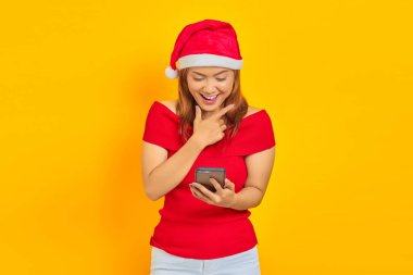 Neşeli genç Asyalı kadın Noel şapkası takıyor ve sarı arka planda parmakları çenesinde olan akıllı telefon kullanıyor.