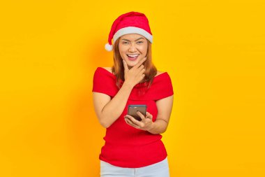 Neşeli genç Asyalı kadın Noel şapkası takıyor ve sarı arka planda parmakları çenesinde olan akıllı telefon kullanıyor.