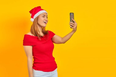 Neşeli genç Asyalı kadın Noel şapkası takıyor sarı arka planda video görüşmesi yapıyor.