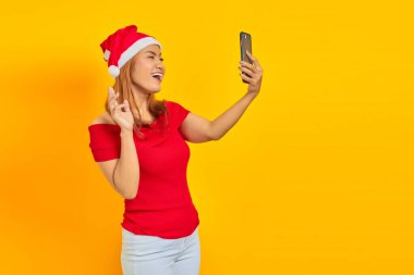Neşeli genç Asyalı kadın Noel şapkası takıyor ve sarı arka planda cep telefonuyla selfie çekiyor.