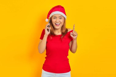 Neşeli genç Asyalı kadın Noel şapkası takıyor cep telefonuyla konuşuyor ve sarı arka planda bir fikirle işaret ediyor.