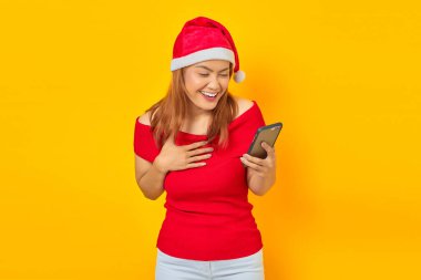 Noel Baba şapkalı heyecanlı genç Asyalı kadın. Elinde cep telefonu, göğsü sarı arka planda.