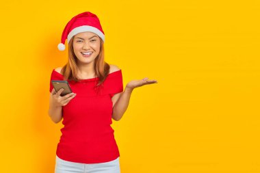 Noel Baba şapkalı gülümseyen Asyalı bir kadının portresi. Akıllı telefon kullanıyor ve sarı arka planda avuç içlerindeki fotokopi alanını gösteriyor.