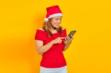 Sarı arka planda izole edilmiş cep telefonu kullanan, Noel Baba şapkalı gülümseyen Asyalı genç kadının portresi.