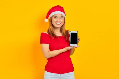 Noel Baba şapkalı gülümseyen genç Asyalı kadın sarı arka planda izole edilmiş boş ekran cep telefonunu gösteriyor.