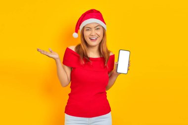 Noel Baba şapkalı gülümseyen genç Asyalı kadın elini uzatıp sarı arka planda akıllı telefonu göstermeye davet ediyor.