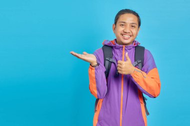 Portrait of smiling young Asian man showing palm hands and thumbs up on blue background