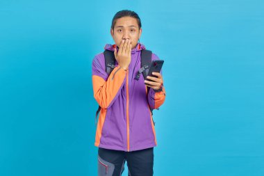 Shocked young handsome man holding smartphone and covering mouth on blue background