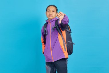 Upset handsome asian man in purple jacket showing thumbs down isolated over blue background