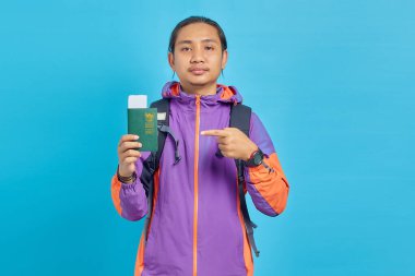 Portrait of cheerful asian young man wearing purple jacket pointing passport book with finger isolated on blue background
