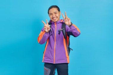 Portrait of smiling young Asian man showing peace sign with fingers and looking at camera isolated on blue background