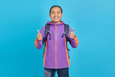 Smiling handsome Asian man in sweater showing thumbs up gesture with hands isolated over blue background