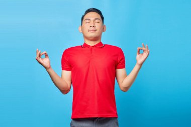 Kırmızı tişörtlü yakışıklı Asyalı genç adam portresi. Mavi arka planda izole edilmiş parmaklarıyla meditasyon hareketi yapıyor ve gözleri kapalı gülümsüyor.