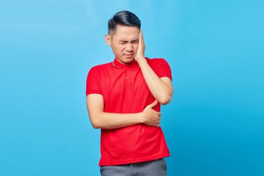 Kırmızı tişörtlü yakışıklı Asyalı genç adamın portresi baş dönmesinden muzdarip, mavi arka planda izole edilmiş baş ağrısı çekiyor.