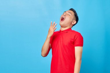 Kırmızı tişörtlü yakışıklı Asyalı genç adamın portresi. Mavi arka planda, ağzı kapalı, uykulu bir hareket sergiliyor.