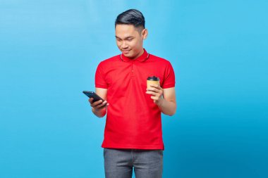 Gülümseyen Asyalı genç adamın portresi cep telefonu kullanıyor ve mavi arka planda kahve tutuyor.