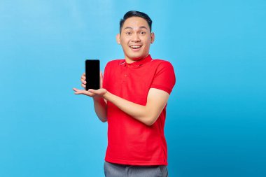 Kırmızı gömlekli neşeli genç Asyalı adamın portresi boş ekran cep telefonu gösteriyor ve mavi arka planda izole edilmiş kameraya bakıyor.