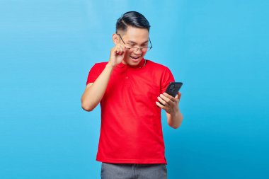 Gözlüklü gülümseyen Asyalı genç adamın portresi ve mavi arka planda izole edilmiş cep telefonundan gelen mesajı okumak.