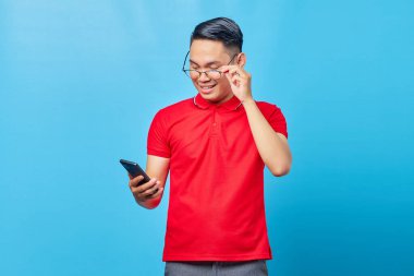 Gözlüklü gülümseyen Asyalı genç adamın portresi ve mavi arka planda izole edilmiş cep telefonundan gelen mesajı okumak.