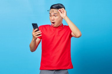 Asyalı genç adamın gözlüklü portresi ve mavi arka planda izole edilmiş akıllı telefondan gelen mesajları okuyor.