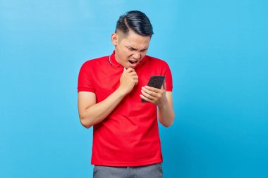 Cep telefonuna bakan, mavi arka planda izole edilmiş sahte haberler okuyan şaşırmış Asyalı genç adamın portresi.