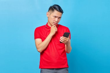 Cep telefonu kullanan ve ciddi görünen düşünceli Asyalı genç adamın portresi mavi arka planda izole edilmiş bir soruyu düşünüyor.