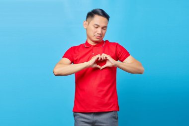 Kırmızı tişörtlü yakışıklı Asyalı adam mavi arka planda kalp hareketi gösteriyor.