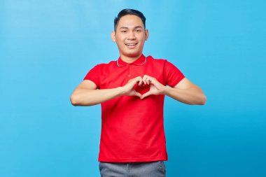 Kırmızı tişörtlü yakışıklı Asyalı adam kalp hareketi gösteriyor ve mavi arka planda izole edilmiş kameraya bakıyor.