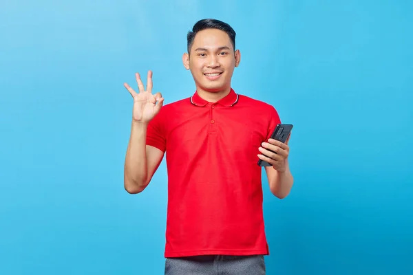 笑顔若いアジアの男の肖像携帯電話を保持し 青い背景に隔離された大丈夫なサインをジェスチャー — ストック写真