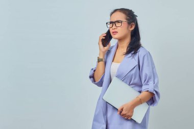 Kafası karışmış, cep telefonuyla konuşan ve dizüstü bilgisayarını beyaz arka planda tutan Asyalı genç kadının portresi