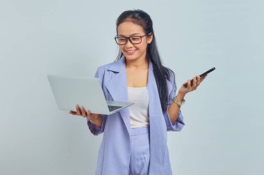 Genç Asyalı kadın portresi dizüstü bilgisayar kullanıyor ve cep telefonunu beyaz arka planda izole edilmiş neşeli bir ifadeyle tutuyor.