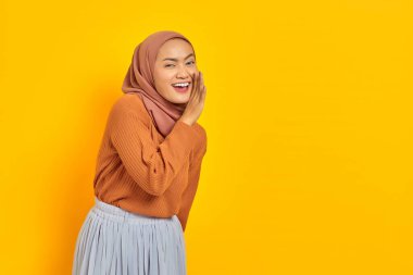 Kahverengi süveter ve el ve ağız tesettürlü güzel bir Asyalı kadın gizli şeyler söylüyor, fısıldıyor, sarı arka planda izole edilmiş neşeli bir ifadeyle kameraya bakıyor. İnsanlar İslam dini kavramları