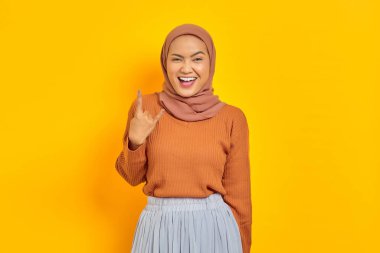 Kahverengi kazaklı ve tesettürlü güzel bir Asyalı kadın çılgın bir ifadeyle bağırıyor. Sarı arka planda elleri izole bir şekilde kaya sembolü yapıyor. İnsanlar İslam dini kavramları