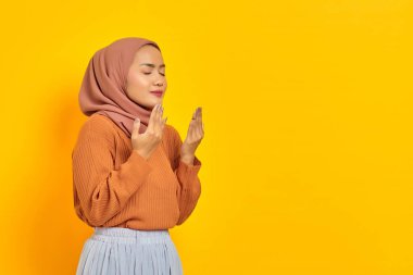 Kahverengi süveter ve tesettürlü güzel bir Asyalı kadın, elleri dua jesti içinde katlanmış, sarı arka planda izole edilmiş bir şey için yalvarıyor. İnsanlar İslam dini kavramları